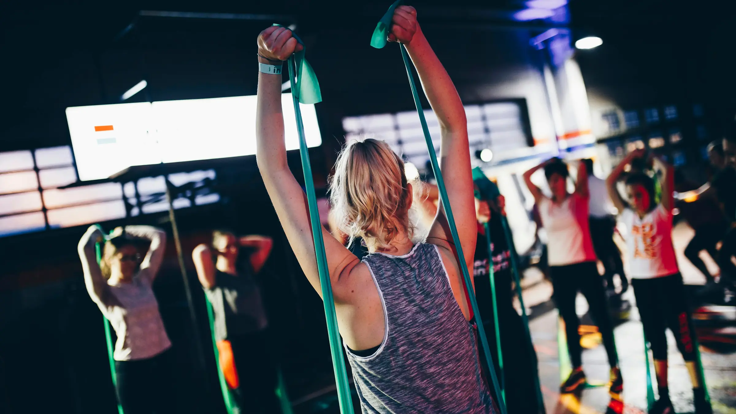 A small group class training with resistance bands, arms overhead, in a dim studio.