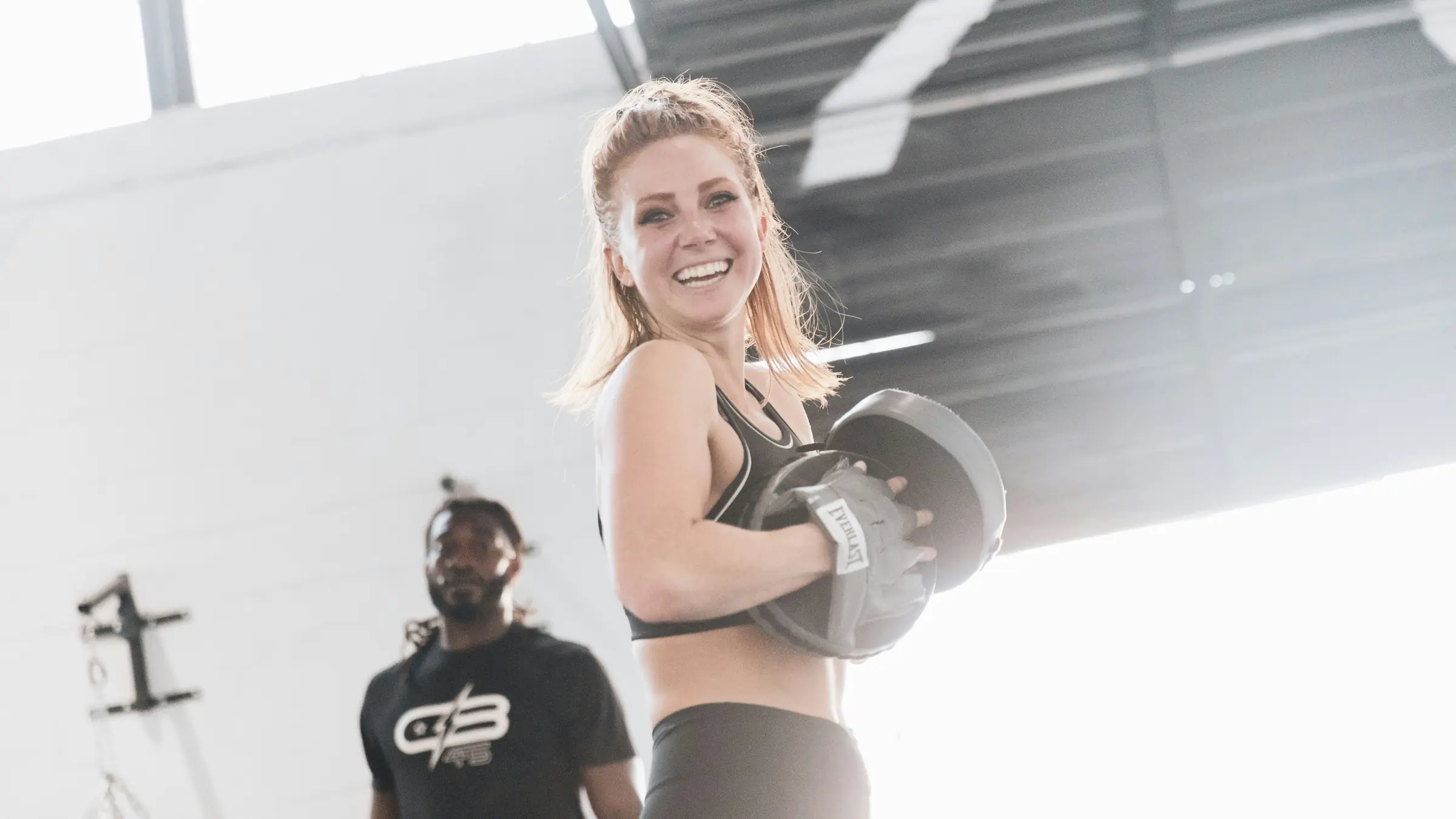 A client laughs mid-set, holding a weight plate, while her coach watches.