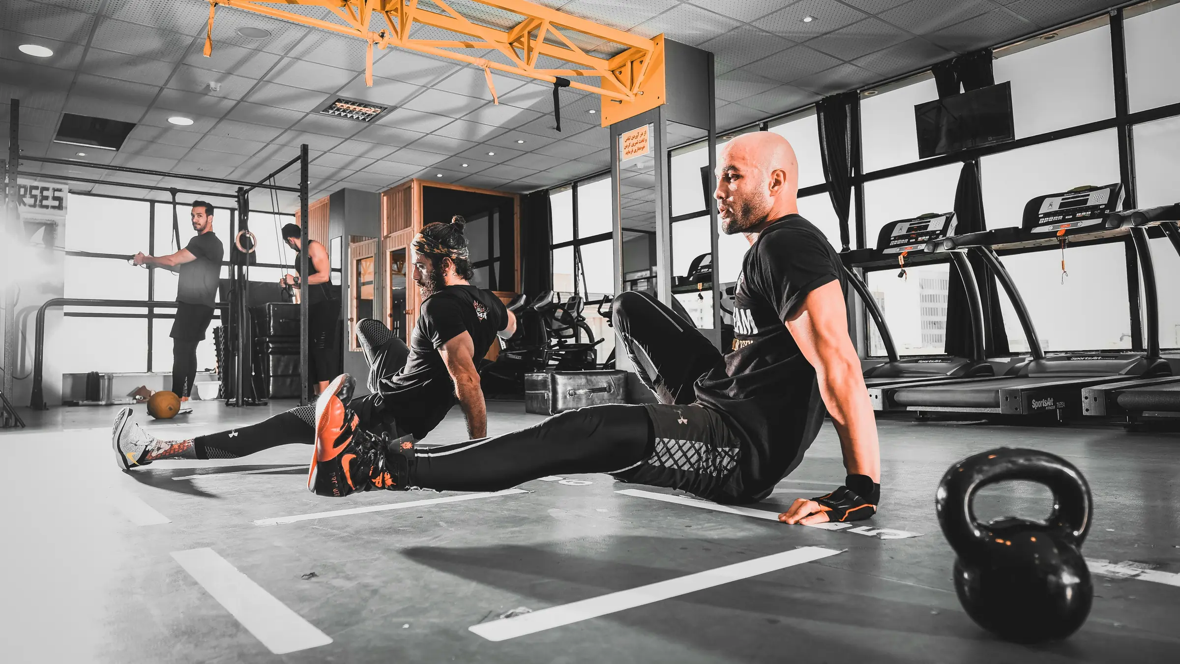 A coach and his client sit on the gym floor between sets, talking, a kettlebell beside them.