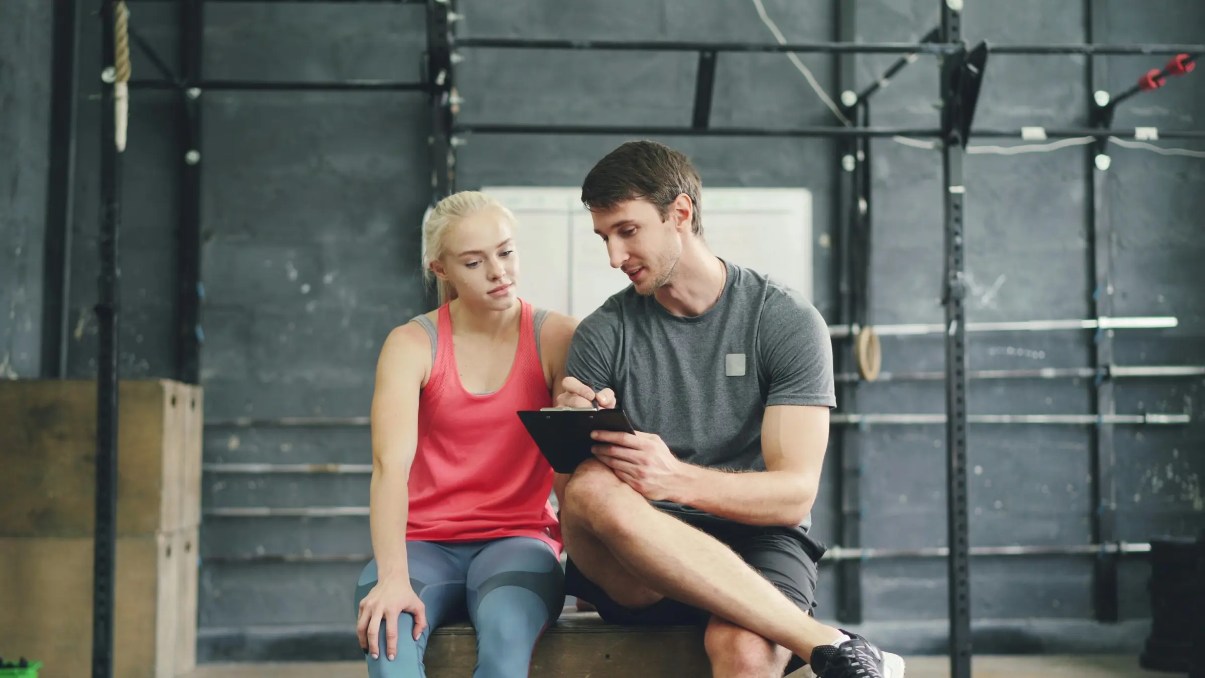A trainer and a client sit side by side on a box in the gym, going over her numbers on his clipboard.