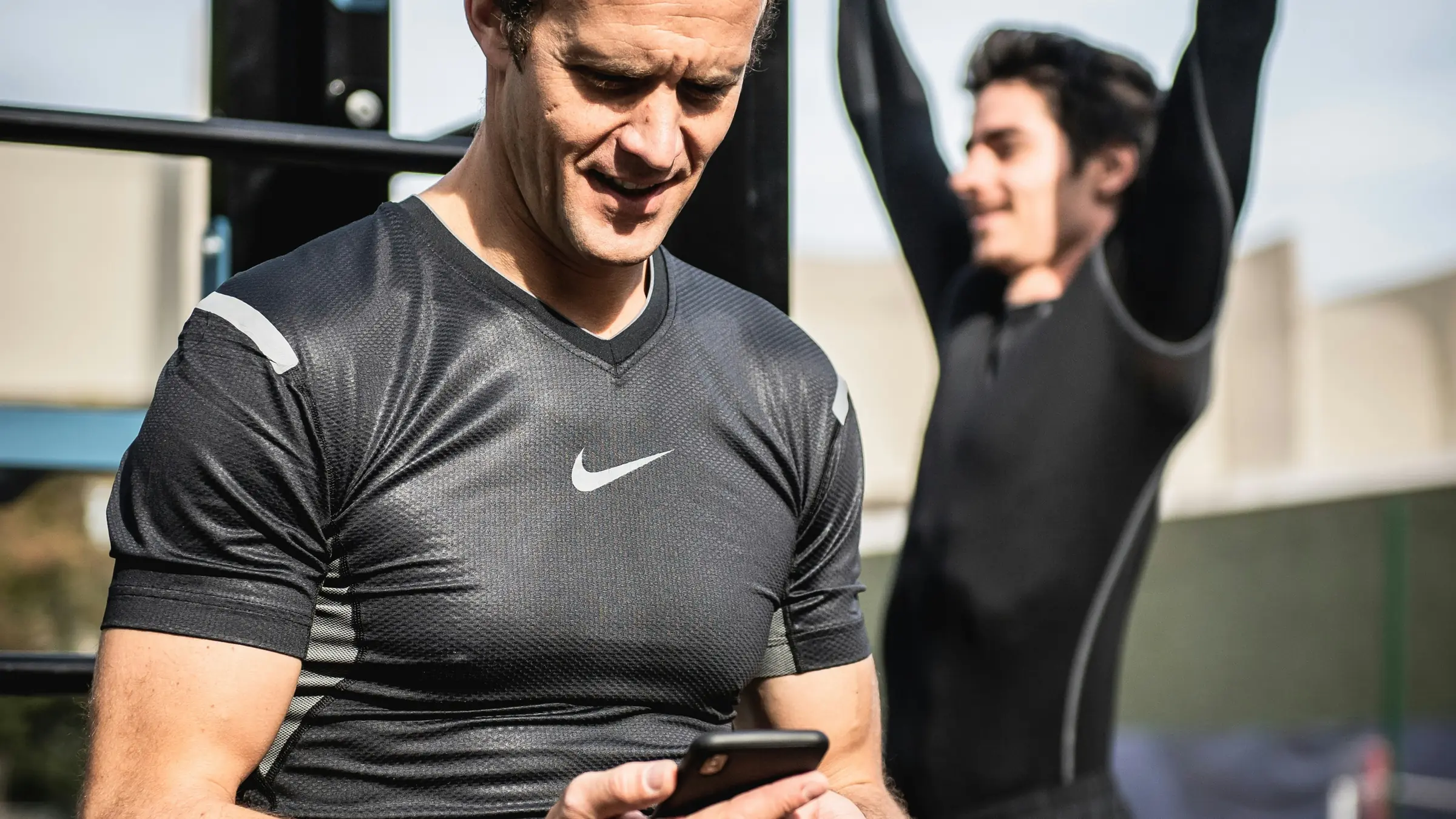 A coach sits by the rig checking his phone while a client does pull-ups behind him.